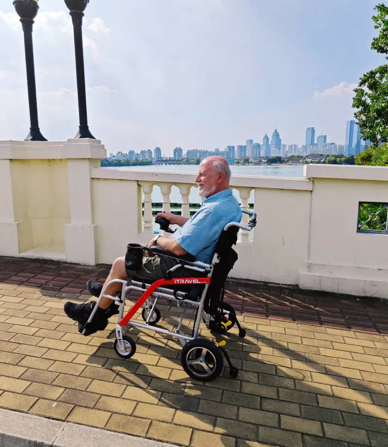 Elderly man sitting on a walker by a riverside with a city skyline in the background.